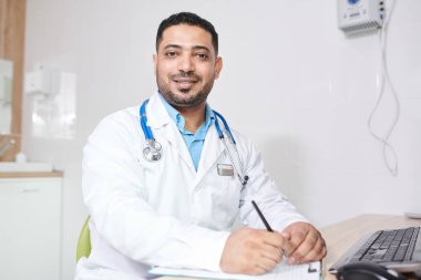 Ofiste masada poz verirken kameraya bakan laboratuvar önlüğü giyen olgun Orta Doğulu doktorun portresi, kopya alanı