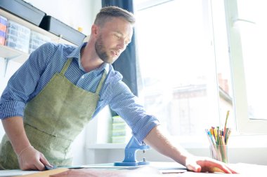 Young serious master in apron working at the table with fabric clipart