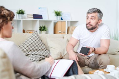 Öfkeli duygusal yakışıklı sakallı adam yastıkları ile kanepede oturan ve psikolog ile acılarını paylaşırken eşleri zina video gösteren