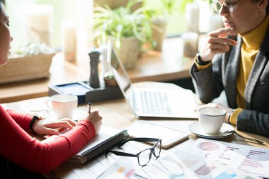 Close-up of pensive businesswomen sitting at table with analytical papers and devices and elaborating business strategy in modern cafe: Asian woman writing plan in notepad clipart