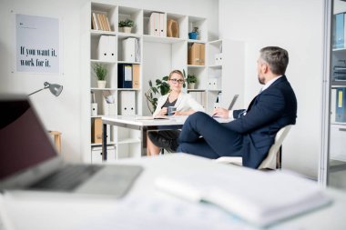 Bir sonraki adımlarını değerlendirirken, kitaplık ve motive pankartile modern ofiste masabaşında oturan ve proje planını tartışan formalgiyimli kendine güvenen işletme yöneticileri