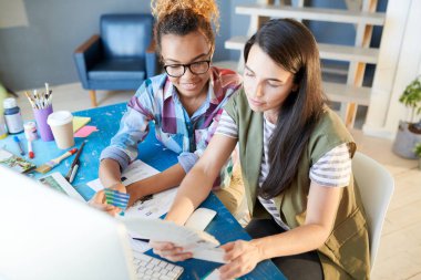 İki yaratıcı genç kadın, biri Afrikalı, güneş ışığı altında masada çalışırken mutlu gülümseyen yüksek açılı portre