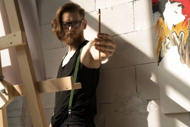 Güneş ışığıyla aydınlatılan sanat stüdyosunda şelonla duran resim sanatına bakan çağdaş uzun saçlı sanatçının bel den yukarı portresi, kopya alanı