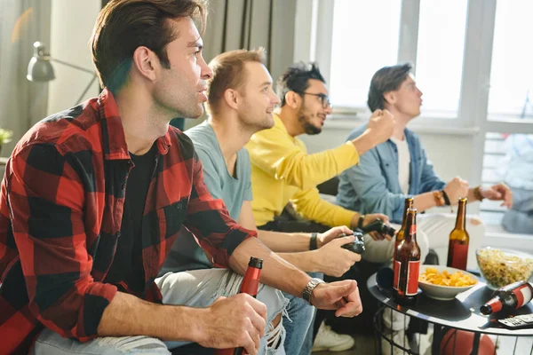 年轻人坐在沙发上喝啤酒 为朋友们在电视上玩电子游戏欢呼 — 图库照片
