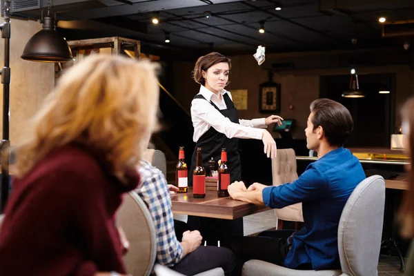 Aggressive displeased attractive young waitress in black apron standing at table and throwing money in guests face while expressing his neglect in dark restaurant