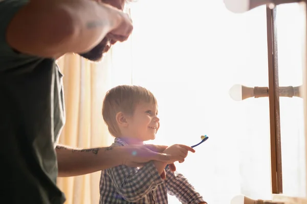 若いです父と正の小さな息子とともにブロンド毛立ちでミラーとクリーニング歯に一緒にバスルーム — ストック写真