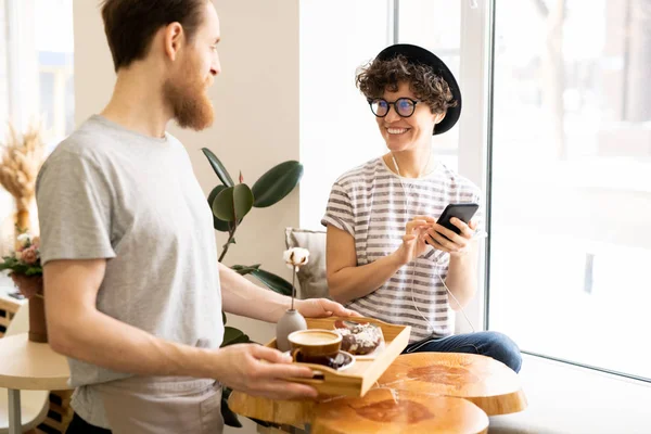 모자와 안경을 창틀에 이어폰으로 음악을 쾌활한 아가씨 고객이 커피와 도넛을 — 스톡 사진