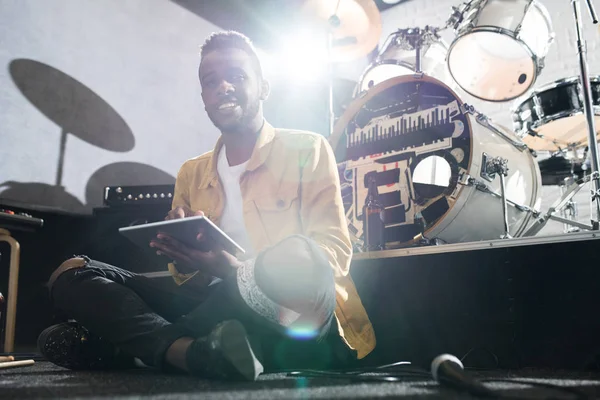 Full length portrait of contemporary  African-American man smiling at camera while sitting on floor using digital tablet during band rehearsal in studio, copy space