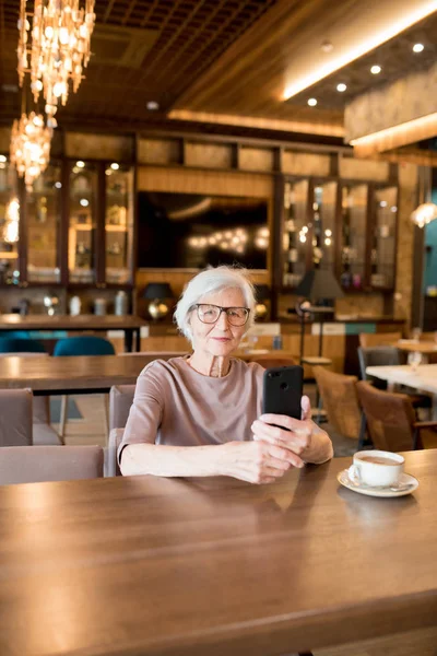 コーヒーカップでテーブルに座り カフェでカメラを見ながらガジェットを使用して メガネでインテリジェントな魅力的な老婦人のコンテンツ — ストック写真