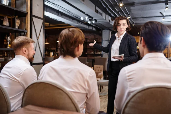 Serious confident attractive young female restaurant manager with short hair standing in front of waiters and using tablet while explaining tasks to staff during briefing