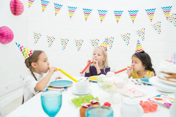 陽気なパーティー帽子でかなり多民族の女の子がカラフルな食器と吹くパーティーの角でテーブルに座って 誕生日に楽しんでいる間に興奮 — ストック写真