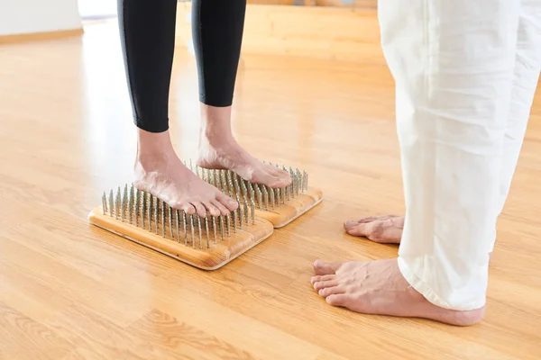Close-up of unrecognizable brave woman in leggings standing barefeet on bed of nails at yoga practice, yoga instructor supporting her