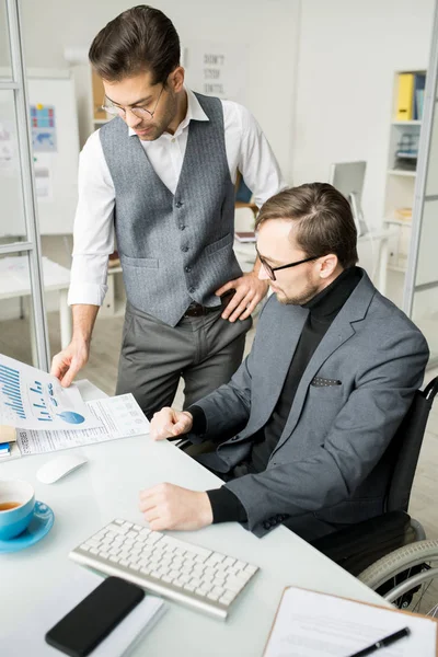 車椅子の障害者の同僚の近くに立っている若いビジネスマンは オフィスで働いている間に財務グラフを一緒に調べています — ストック写真