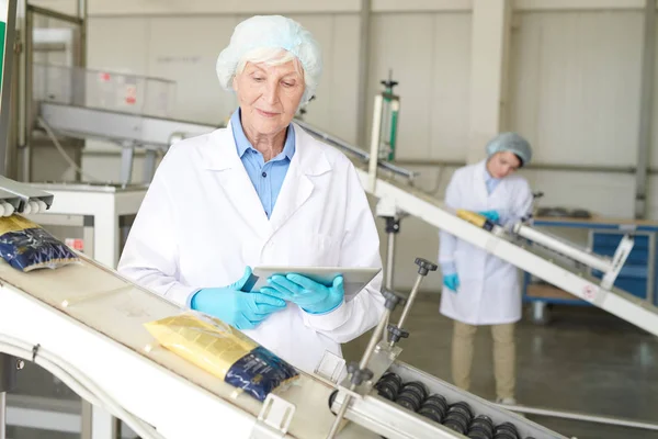 現代の食品工場で生産プロセスを制御し コンベアベルトを滑り落ちるマカロニバッグを見て シニア女性の肖像画 コピースペース — ストック写真