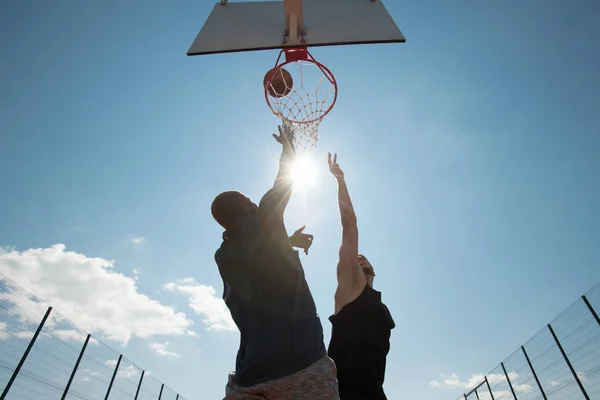 Low angle view of two young men playing basketball and jumping by hoop against blue sky, copy space