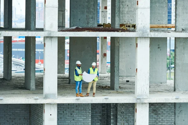 Dwóch Mężczyzn Kaskach Kamizelkach Czytanie Schemat Budowy Stojąc Betonowej Podłodze — Zdjęcie stockowe