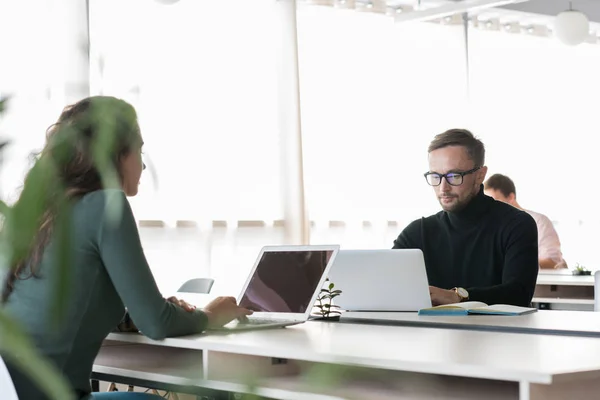 Busy office workers using portable computers in modern open space: concentrated handsome businessman in glasses sitting at table and analyzing data