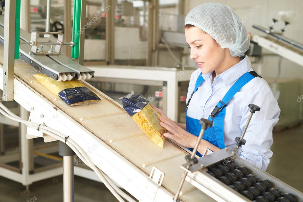 Portrait of female factory worker controlling  packaging process at modern food factory and looking at macaroni bags sliding down conveyor belt, copy space