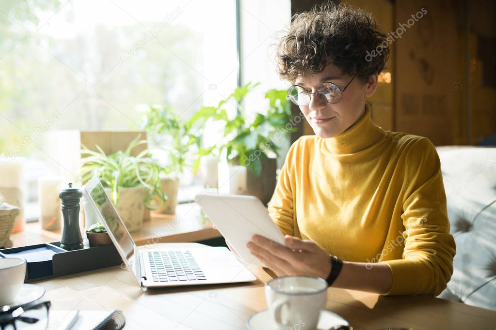 Content pensive curly-haired female freelancer in glasses sitting at table in modern cafe and using digital tablet while analyzing online information