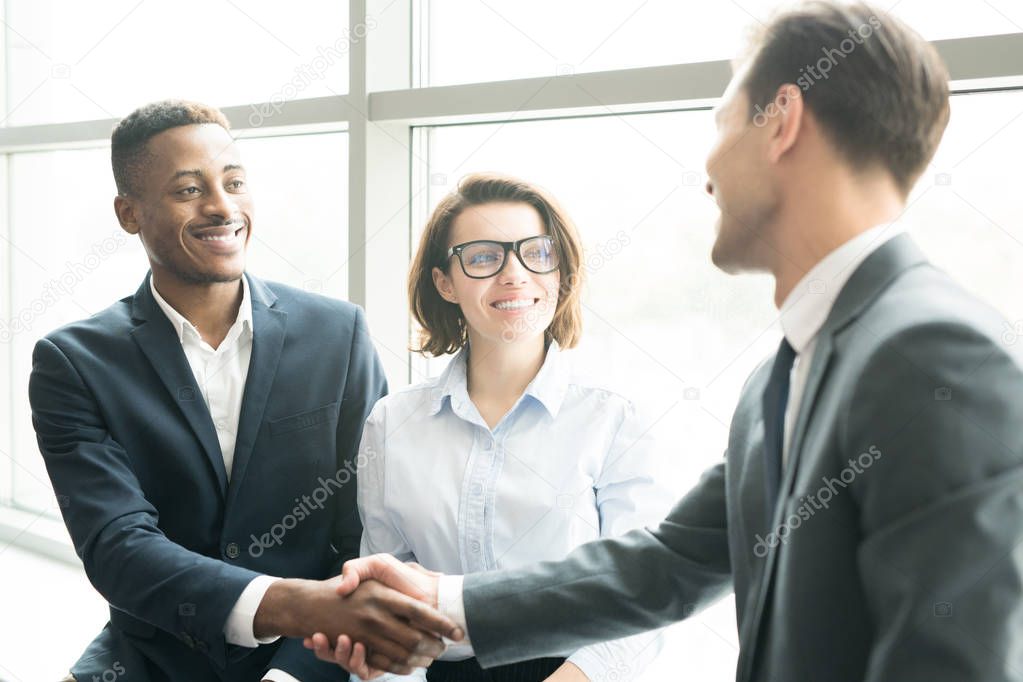 Cheerful successful business managers shaking hands of African-American businessman while congratulating young specialists with concluding contract