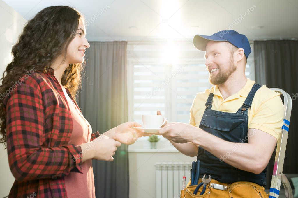 Smiling hospitable attractive lady with curly hair giving coffee to happy bearded repairman in cap sitting on stepladder