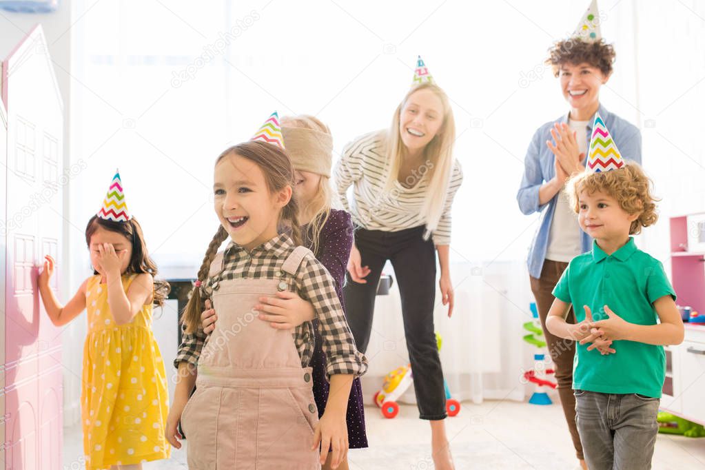 Positive blindfolded girl in cute dress embracing excited friend in party hat while capturing her during game at birthday party