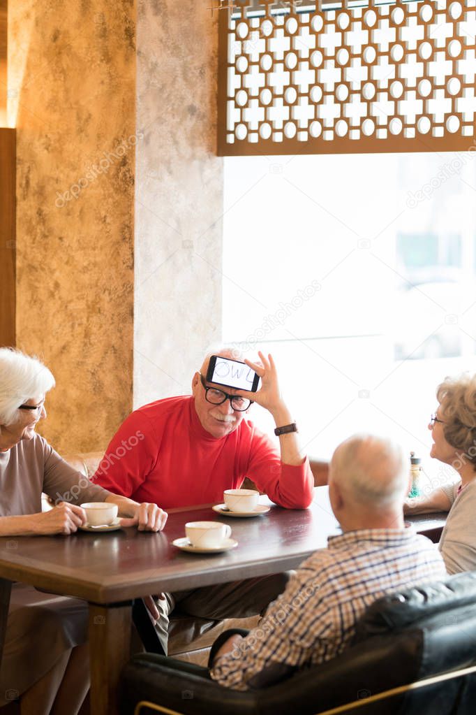 Puzzled handsome senior man holding smartphone with word on display while playing charades with friends in modern cafe