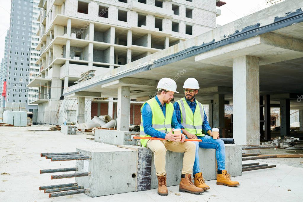 Serious content young multi-ethnic construction coworkers sitting on concrete beam with steel bars and chatting while drinking coffee during break