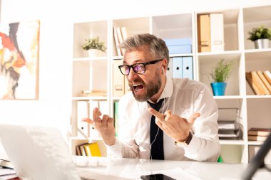 Annoyed furious bearded businessman in glasses and earphones sitting at office table and showing fucks to laptop while knowing bad news