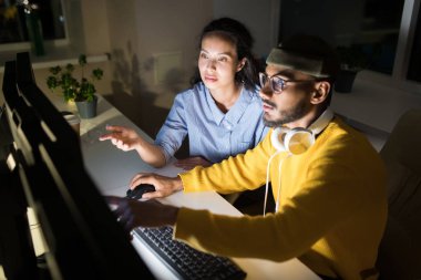 Gece geç saatlerde karanlık ofiste bilgisayar kullanırken ekrana işaret eden ve projeyi tartışan iki meslektaşının portresi