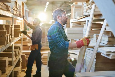 Güvenlik gözlüğü ve iş eldivenleri giyen sakallı ciddi gri saçlı el işçisi raflarda ayakta ve depoda ahşap plakalar kontrol