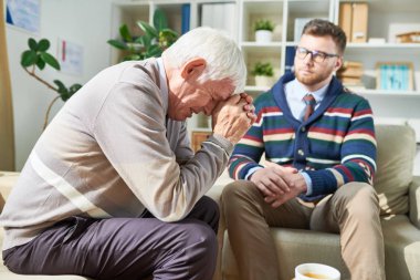 Psikoterapi seansı sırasında ağlarken kanepede oturan ve ellerine yaslanan üzgün duygusal kıdemli hasta, onu dinleyen özenli psikiyatrist