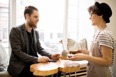 Şapka ve önlük pozitif çekici garson müşteriye kahve ve vazo ile tepsi veren, kulaklık neşeli sakallı adam ahşap masada oturan ve garson gülümseyen