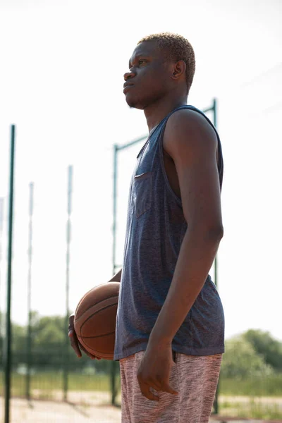 Side view portrait of modern African-American man standing in basketball court holding ball and looking away