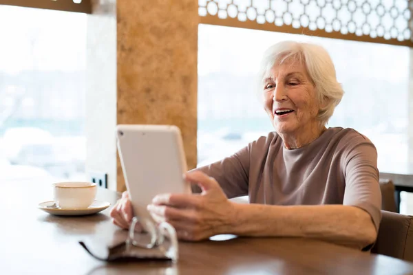陽気な興奮した白髪の先輩女性が木製のテーブルに座り デジタルテーブルでビデオを見ながら笑いながら 彼女はカフェでリラックス — ストック写真