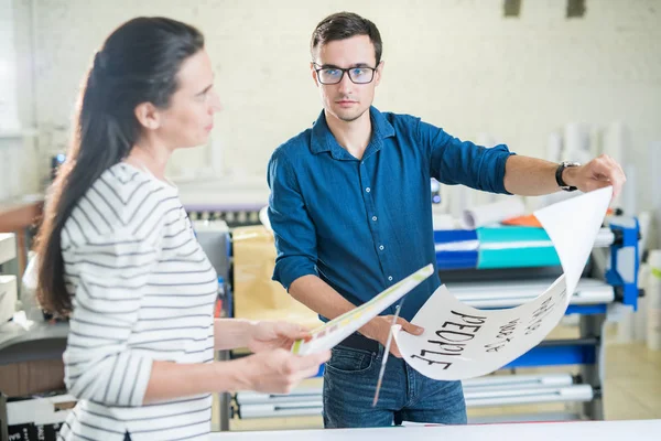 Young woman and man in office of publishing house discussing paper placard with printed text