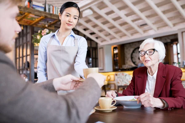 若いウェイトレスがレストランのテーブルに座っている間に若い男性と高齢者の女性にコーヒーを持ち込みます — ストック写真