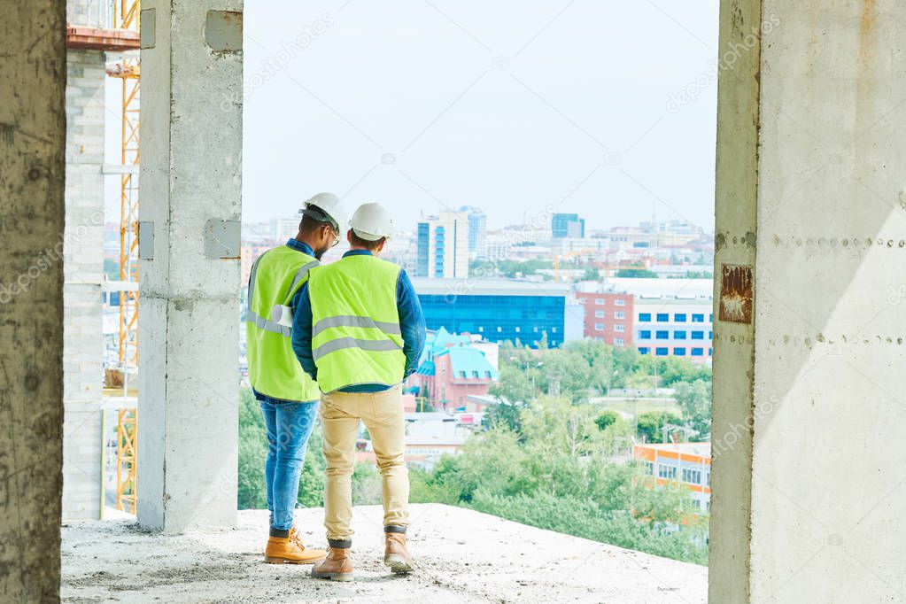Serious busy men in hardhats and green vests working at construction site of apartment building and discussing infrastructure while contemplating city