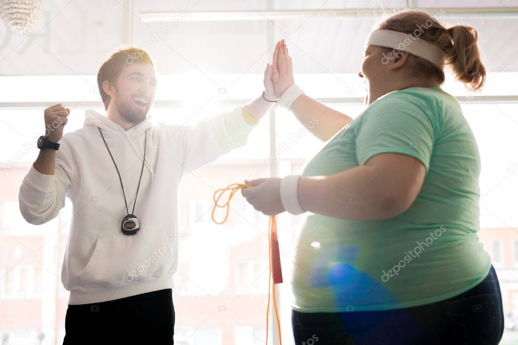 Waist up portrait of smiling fitness coach high fiving with fat young woman during workout in sunlight