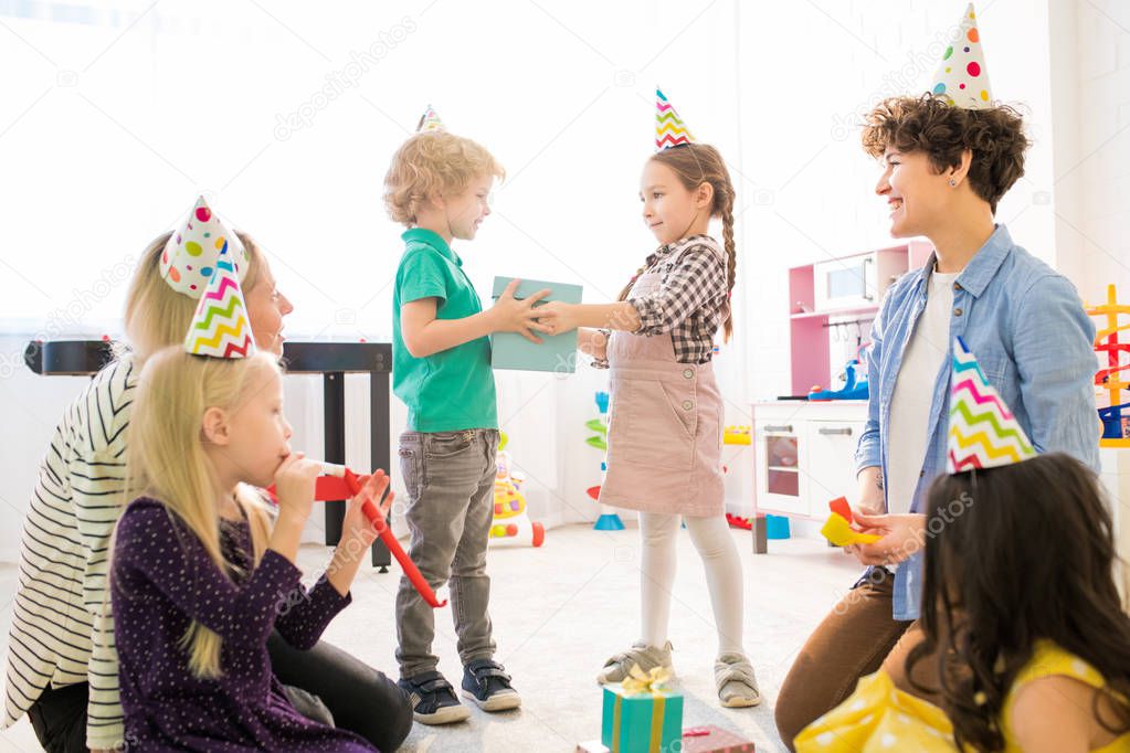 Time for gifts at birthday party: positive cute girl with braids and giving gift box to curly-haired boy while congratulating him with birthday at party