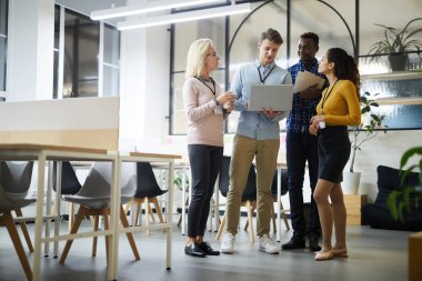 Proje yi tartışırken, ortak çalışma alanında duran ve dizüstü bilgisayardaki çevrimiçi verileri analiz eden gündelik giyimli kendinden emin çok ırklı meslektaşlarından oluşan bir grup
