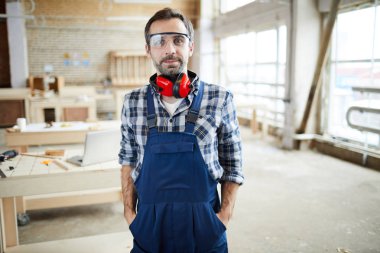 Tulum giyen ve kulak koruyucular ceplerinde elele ve kamera atölye bakarak boyunda yakışıklı orta yaşlı marangoz güvenlik gözlüğü içerik memnun.