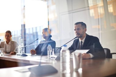 Konferansta meslektaşları arasında oturan ve mikrofon hoparlörle konuşan takım elbiseli ciddi delege