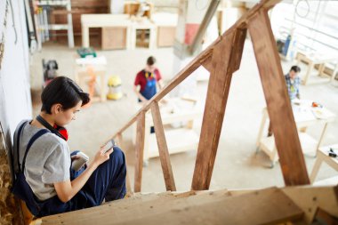 Kısa saç tulum giyen ve kulak koruyucular atölyesinde kısa bir mola yaparken merdiven ve smartphone cep telefonu ile mesaj sms oturan boynunda ciddi dalgın genç bayan marangoz.