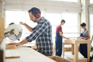 Kulak koruyucular ve emanet ciddi konsantre orta yaşlı marangoz tezgah ayakta ve atölyede ahşap tahta kesmek için daire testere kullanarak koruyucu gözlük