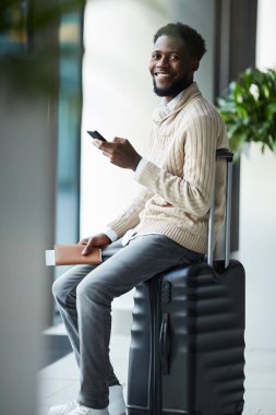 Hafif bej kazak ve uçuş duyurusu için beklerken bavula oturan gri kot neşeli genç Gezgin