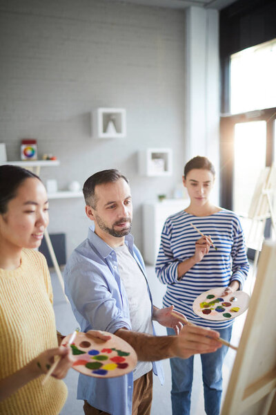 Two girls with palettes and paintbrushes looking at easel with their teacher work at lesson in studio