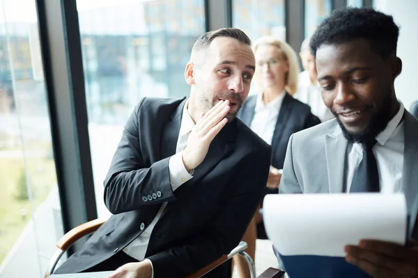 Young businessman whispering something to colleague sitting next to him while listening to report of speaker at conference