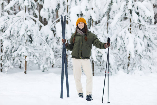 Full length portrait on active young woman holding skis standing in winter forest and smiling happily at camera, copy space