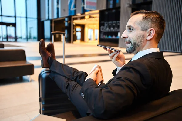 Young mobile businessman using voice messenger while recording message on smartphone in airport lounge